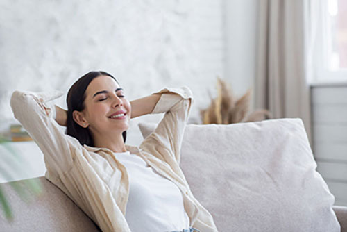 A woman with long hair leans back on a sofa with her hands behind her head, smiling and appearing relaxed in a well-lit room.