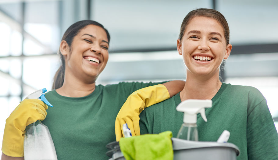 Two women in green shirts laughing while holding cleaning supplies and wearing yellow gloves.